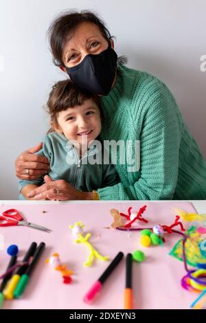 Grand-mère embrassant la petite-fille souriante à table à la maison pendant une pandémie Banque D'Images