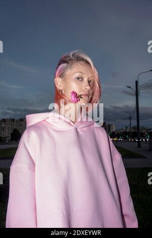 Portrait d'une jeune femme aux cheveux roses portant une chemise à capuchon rose tenant la rose dans la bouche Banque D'Images