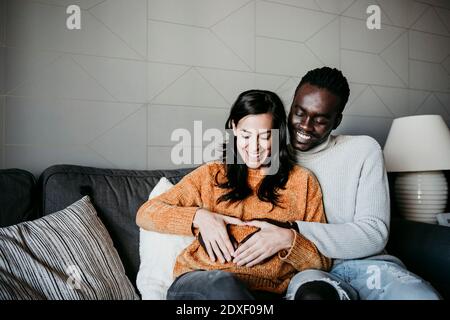 Homme heureux et femme enceinte formant le coeur avec les mains sur le ventre tout en étant assis sur un canapé à la maison Banque D'Images