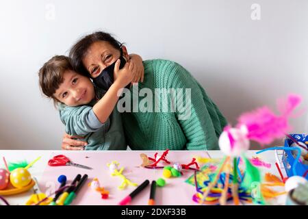 Petite-fille embrassant la grand-mère à la table contre le mur à la maison pendant COVID-19 Banque D'Images