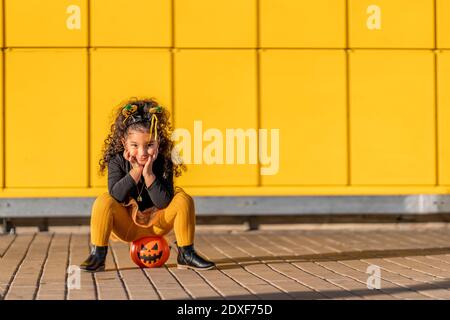 Fille assise avec la tête dans les mains sur le pot de fleur d'Halloween par rapport à la paroi à motif coché jaune Banque D'Images