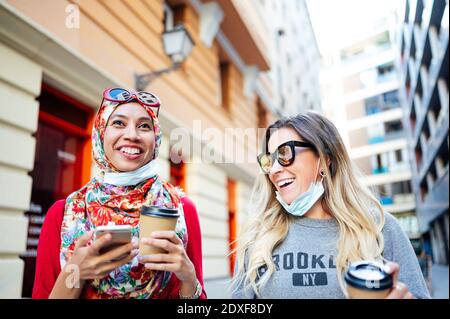 Des amies joyeuses rient tout en tenant des tasses jetables en ville Pendant COVID-19 Banque D'Images