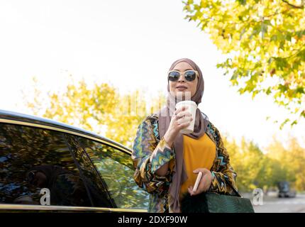 Jeune femme portant le hijab et des lunettes de soleil buvant du café en position debout en voiture Banque D'Images