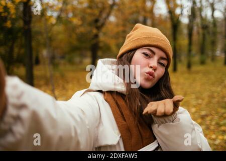 Belle jeune femme soufflant baiser tout en prenant selfie en automne stationnement Banque D'Images
