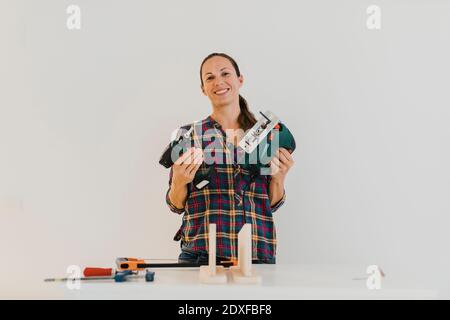 Femme souriante tenant une scie sauteuse électrique et une perceuse tout en se tenant contre mur blanc à la maison Banque D'Images