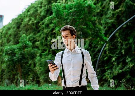 Homme d'affaires portant une chemise blanche et des bretelles noires à la recherche d'une élégante téléphone à l'extérieur Banque D'Images