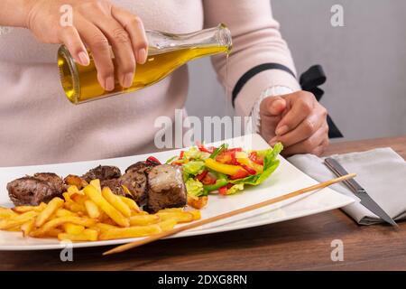 vue d'une femme qui verse de l'huile d'olive sur des légumes fraîchement coupés sur une assiette avec des morceaux de bœuf juteux et des frites. Mise au point sélective. Banque D'Images