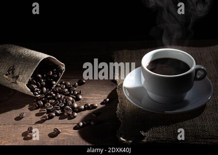 Tasse de café blanc, grains de café déversés du sac sur une table en bois sur fond noir. Banque D'Images