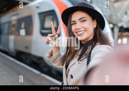 Jolie fille touriste prend selfie à la gare de chemin de fer contre le fond de la locomotive. Une fille parle lors d'un appel vidéo Banque D'Images