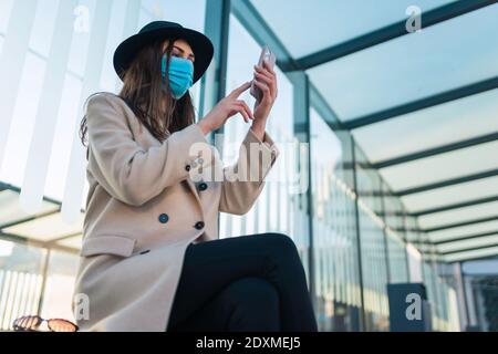 Fille dans le masque de protection est assis à l'arrêt de bus avec le téléphone dans les mains. La femme est assise à l'arrêt d'autobus pendant une pandémie et attend le transport Banque D'Images