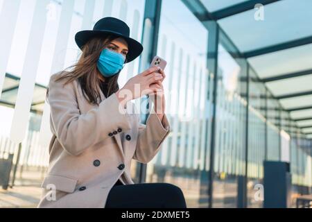 Fille dans le masque de protection est assis à l'arrêt de bus avec le téléphone dans les mains. La femme est assise à l'arrêt d'autobus pendant une pandémie et attend le transport Banque D'Images