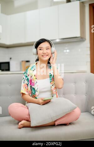 Portrait belle jeune femme asiatique utiliser smartphone mobile avec casque pour écouter de la musique sur le canapé dans le salon Banque D'Images