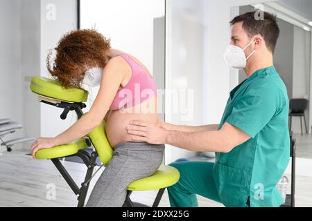 Prise de vue d'un jeune physiothérapeute massant le dos d'une femme enceinte. Homme portant un masque de protection pendant la nouvelle normale. Photo de haute qualité Banque D'Images