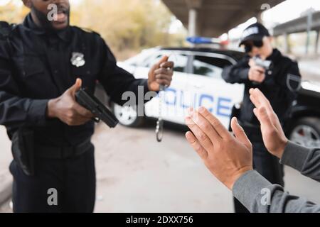 délinquant montrant les mains tandis que des policiers multiculturels tiennent des pistolets et menottes sur fond flou en extérieur Banque D'Images