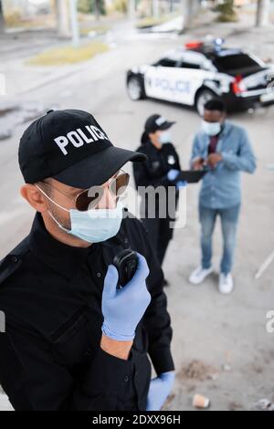 Policier en gants de latex et masque médical avec talkie-walkie alors que l'un de nos collègues parle avec une victime afro-américaine sur un fond flou sur urban str Banque D'Images