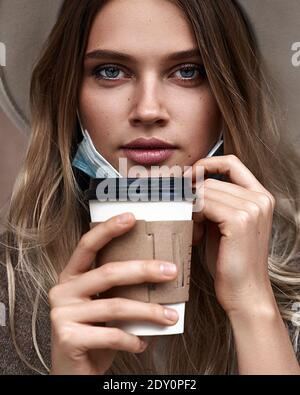 Fin du verrouillage de quarantaine. Une fille vaccinée boit du café tout en tirant sur le masque de protection Banque D'Images