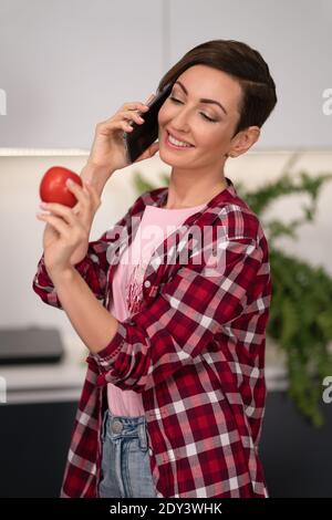 Choisir de la tomate fraîche tout en parlant au téléphone femme au foyer avec une courte coiffure. Déjeuner de cuisine pour la famille dans la cuisine moderne. Alimentation saine à Banque D'Images