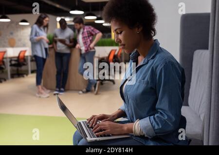 Vue latérale d'une femme afro-américaine dans des vêtements décontractés sur le canapé et en tapant sur le clavier de l'ordinateur portable tout en travaillant projet à proximité de divers collègues Banque D'Images