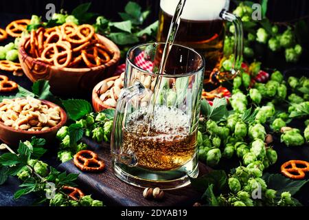 Bière allemande légère versée dans un grand verre, houblon vert frais et bols avec des collations salées et des noix, concept de festival de la bière d'automne, fond sombre, selecti Banque D'Images