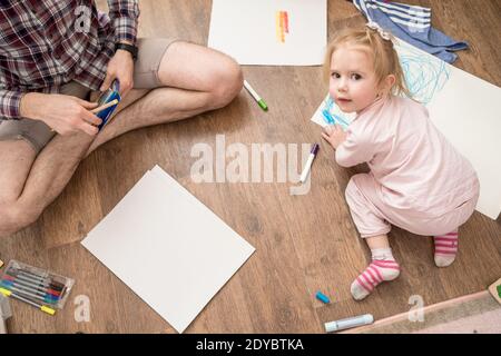 Mignon, heureux enfant dessine avec des marqueurs de couleur sur papier blanc, assis sur le sol, à côté de son père. Style de vie. Banque D'Images