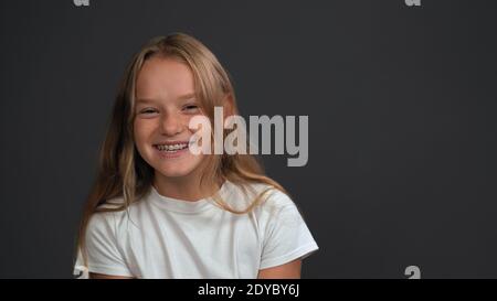 Bonne petite fille souriante avec des cheveux blonds debout regardant l'appareil photo porte un t-shirt blanc isolé sur le gris foncé ou arrière-plan noir Banque D'Images