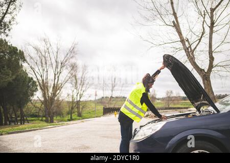 Jeune femme caucasienne regardant le moteur de voiture, voiture en panne au milieu de la route. Concept d'assistance routière et automobile. Banque D'Images