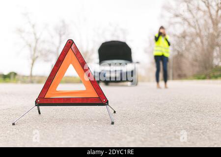 Jeune femme caucasienne, parlant sur son téléphone cellulaire alors que sa voiture est en panne sur la route avec des triangles d'avertissement réfléchissants. Mise au point sélective. Auto Banque D'Images