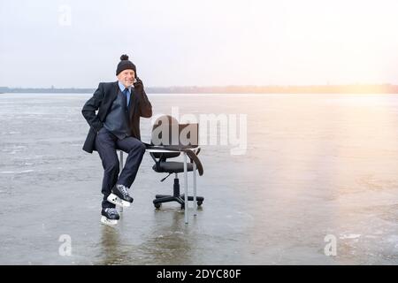 homme d'affaires souriant en costume et patins assis sur la table sur un lac gelé Banque D'Images