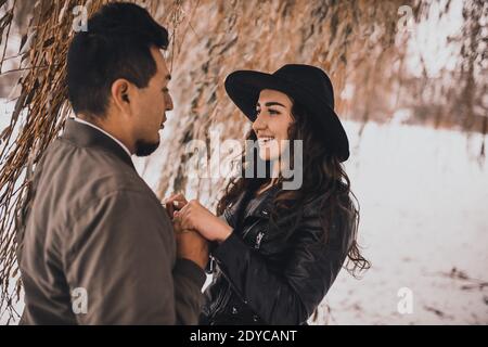 L'homme mexicain tient les mains d'une femme ukrainienne dans une veste en cuir noir et un chapeau noir sur la nature en hiver. Amour entre les différentes nationalités Ven Banque D'Images