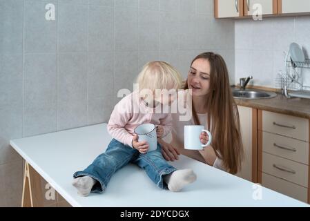 Un enfant curieux regarde dans le mug de la mère tout en étant assis dans la cuisine. Petit déjeuner familial. Banque D'Images