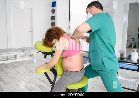Prise de vue d'un jeune physiothérapeute massant le dos d'une femme enceinte. Homme portant un masque de protection pendant la nouvelle normale. Photo de haute qualité Banque D'Images