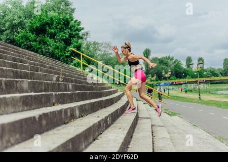Faites de la bonne santé à l'athlète, belle femme dans des vêtements de sport serrés sautant dans les escaliers, en vous réchauffant avant de courir tout en regardant très motivé. Banque D'Images