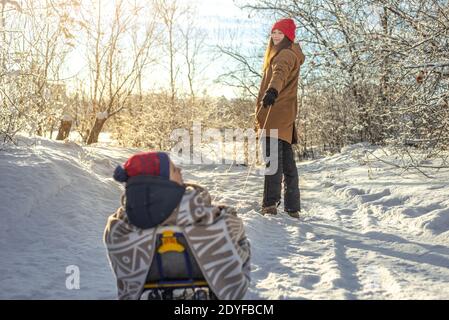 Une mère avec un enfant marche un matin d'hiver glacial le jour du soleil. Bébé enveloppé dans une couverture sur un traîneau Banque D'Images