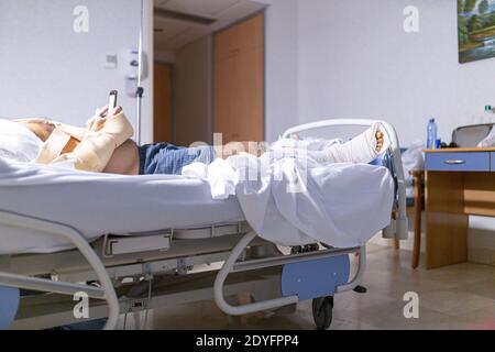 Patient allongé dans le lit de l'hôpital avec une jambe cassée regardant le téléphone cellulaire. Concept d'hospitalisation et de soins médicaux. Banque D'Images