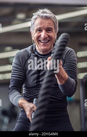 Un homme plus âgé rit tout en s'entraîner avec des cordes à l'intérieur de la salle de gym, en portant des écouteurs. Banque D'Images