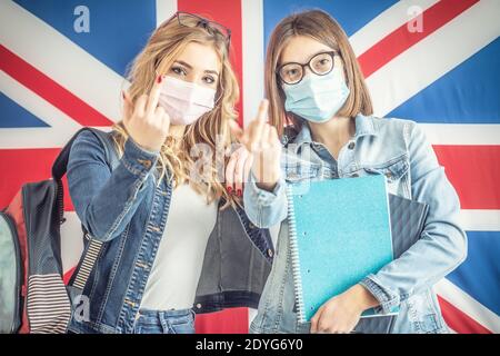 Deux étudiantes portant un masque facial protecteur montrent un geste du doigt central en se tenant devant le drapeau britannique. Banque D'Images