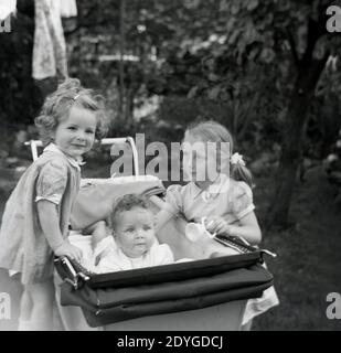 Années 1950, historique, l'été et à l'extérieur dans un jardin, deux petites filles douces, sœurs, debout à côté d'un entraîneur-construit pram ou de la voiture de bébé de l'époque, avec l'une de la fille debout sur la roue et l'autre avec sa main sur leur bébé frère couché dans le pram, Angleterre, Royaume-Uni. Banque D'Images