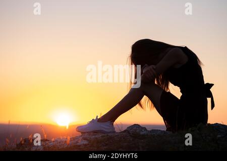 Jeune femme dépressive noire robe courte d'été assise sur une colline de montagne pensant à l'extérieur au coucher du soleil. Femme solitaire contemplant dans une soirée chaude Banque D'Images