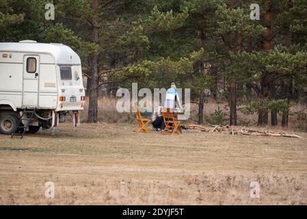 Couple commençant un feu de camp pour avoir le dîner près de leur remorque sur l'herbe dorée en automne forêt de pins verts Banque D'Images