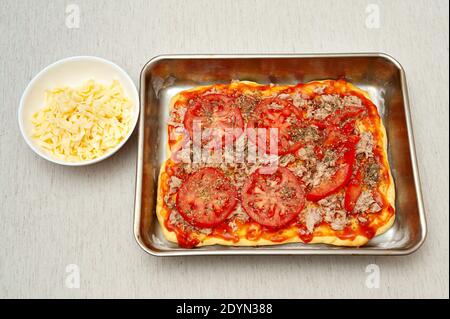 Pizza rectangulaire maison - pâte à pizza avec tomates en tranches, thon et sauce tomate à côté d'un bol de fromage râpé. Vue de dessus. Banque D'Images