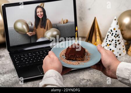 Les mains de sexe masculin tiennent un gâteau avec une bougie devant l'écran de l'ordinateur portable. Jeune femme célébrant son anniversaire à la maison par appel vidéo en ligne avec Banque D'Images