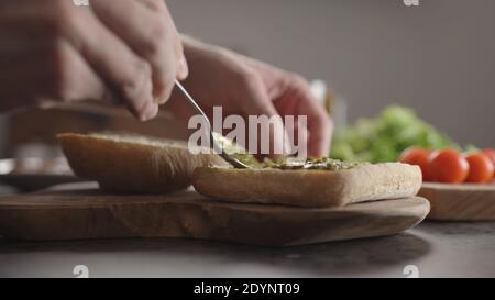 Homme étalant du pesto sur un petit pain ciabatta pour un sandwich sur un comptoir de cuisine, photo large Banque D'Images