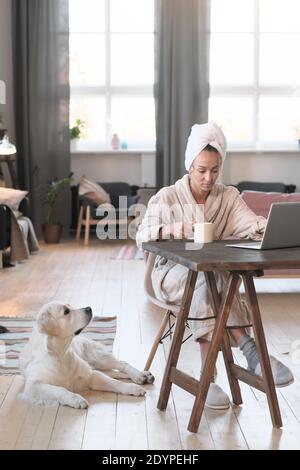 Jeune femme en peignoir assise à la table et travaillant en ligne avec un ordinateur portable à la maison Banque D'Images