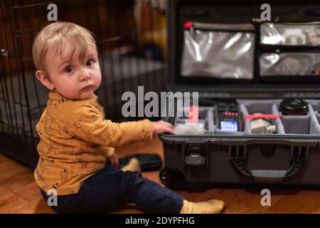 Petit garçon mignon regardant l'appareil photo tout en touchant la valise de son père remplie d'équipement vidéo professionnel. Notion de curiosité et de découverte. Banque D'Images