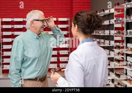 Homme senior regardant de nouvelles lunettes en exposition en se tenant debout devant une jeune ophtalmologiste féminine et en consultation avec elle dans le bureau médical Banque D'Images