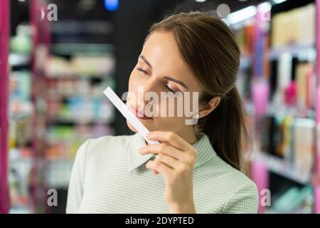 Jeune jolie femme dans une robe élégante testant le parfum tout en tenant le papier buvard par son nez, elle choisit une nouvelle odeur pour elle-même dans la boutique de beauté Banque D'Images