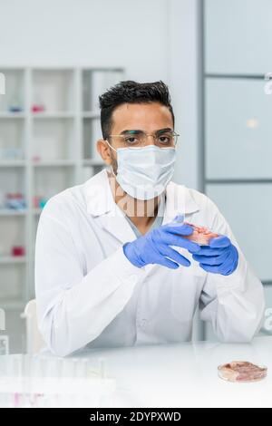 Jeune homme ganté chercheur scientifique en whitecoat, masque et lunettes tenant un morceau de viande végétale brute dans un plat de pétri tout en travaillant en laboratoire Banque D'Images