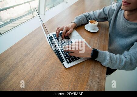 Les mains des hommes sur le clavier de l'ordinateur portable moderne. Concept freelance. Concept de programmateur. Couper la vue du jeune homme travaillant sur l'ordinateur près de la fenêtre avec une tasse de Banque D'Images