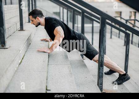 Un jeune homme bien construit qui se relève des escaliers. Vue latérale. Homme de sport aux cheveux noirs portant des vêtements de sport noirs et des baskets qui s'efforcent de faire de l'exercice dans la rue Banque D'Images