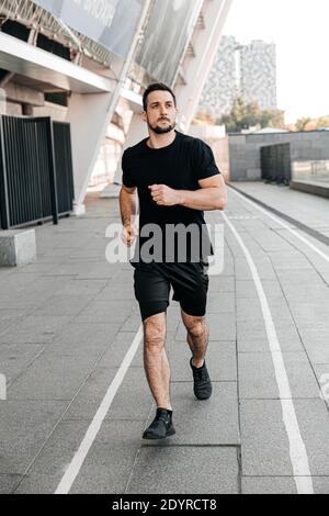 Un homme sportif qui fait du jogging dans la ville. Un mode de vie sain. Homme en train de courir. Style de vie urbain. Vie active. Fitness en plein air. Vêtements de sport noirs. Joyeux homme Banque D'Images